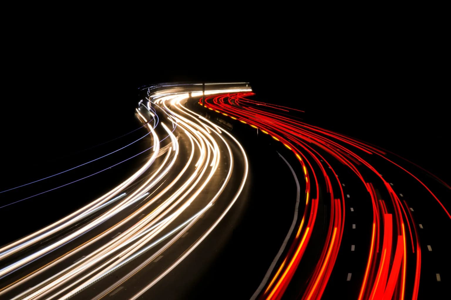 Long-exposure white and red light trails curve through a dark roadway.